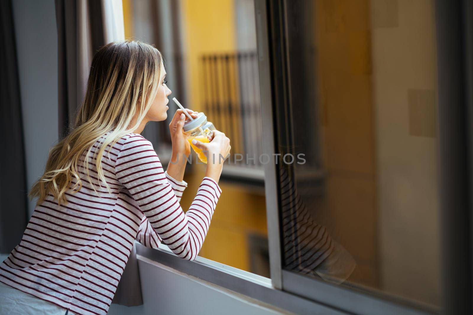 Young woman drinking a glass of natural orange juice, leaning out the window of her home. by javiindy