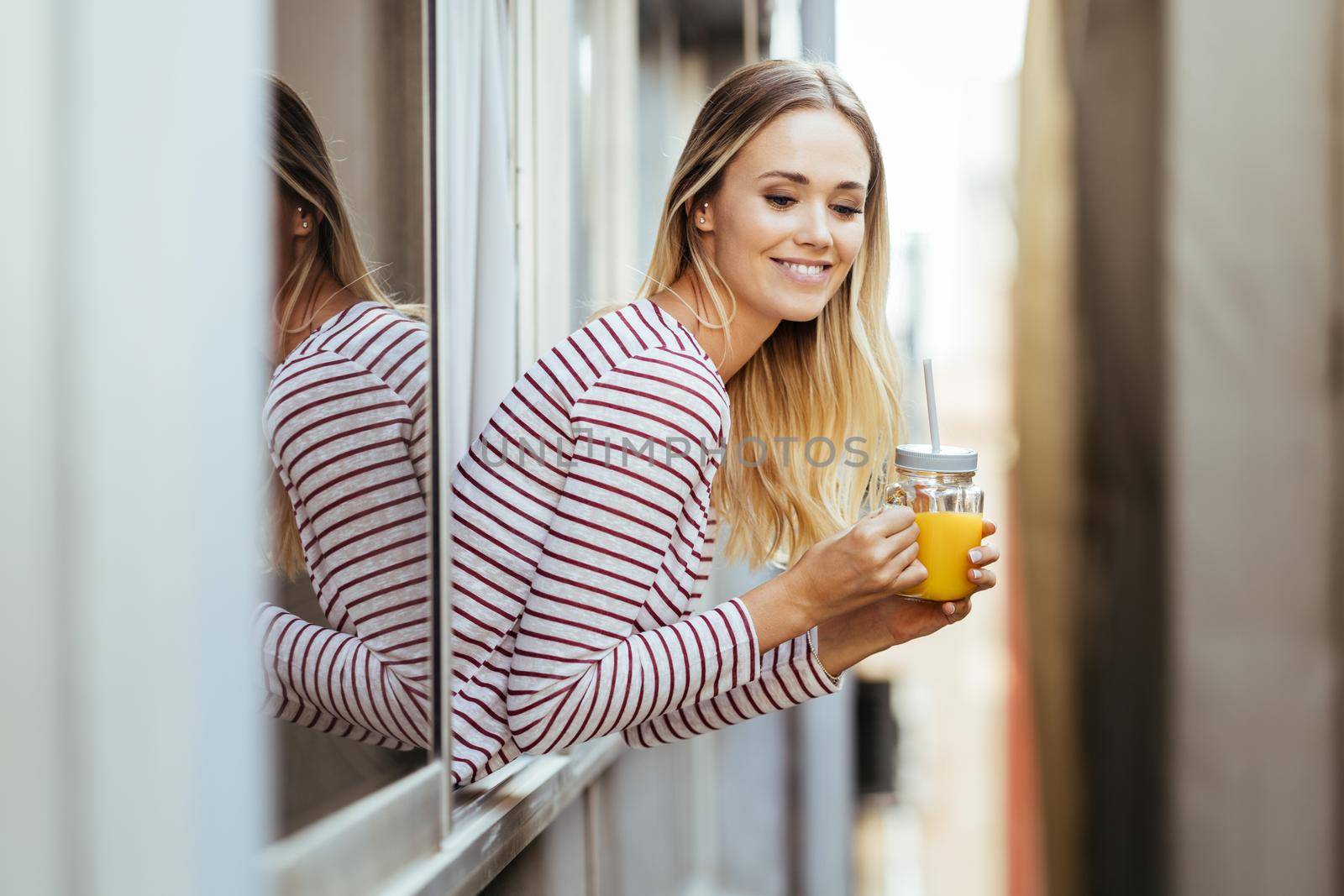 Smiling woman drinking a glass of natural orange juice, leaning out the window of her home. by javiindy