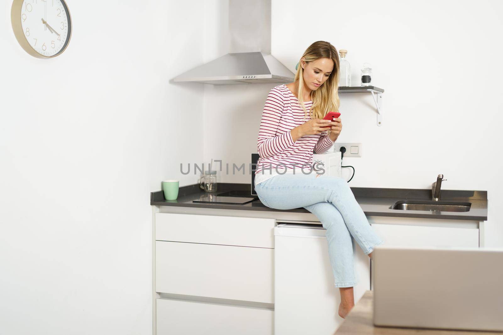 Young woman using her smartphone sitting in the kitchen at home