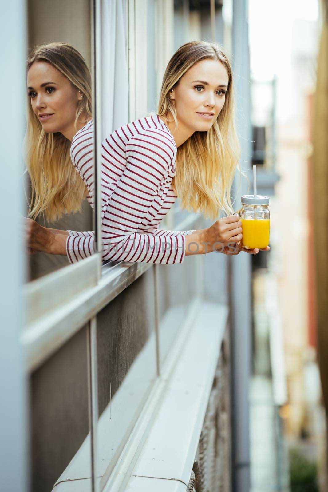Young blonde woman drinking a glass of natural orange juice, leaning out the window of her home.