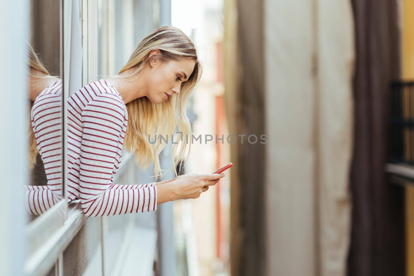 Attractive woman leaning out of her house window using a smartphone. by javiindy