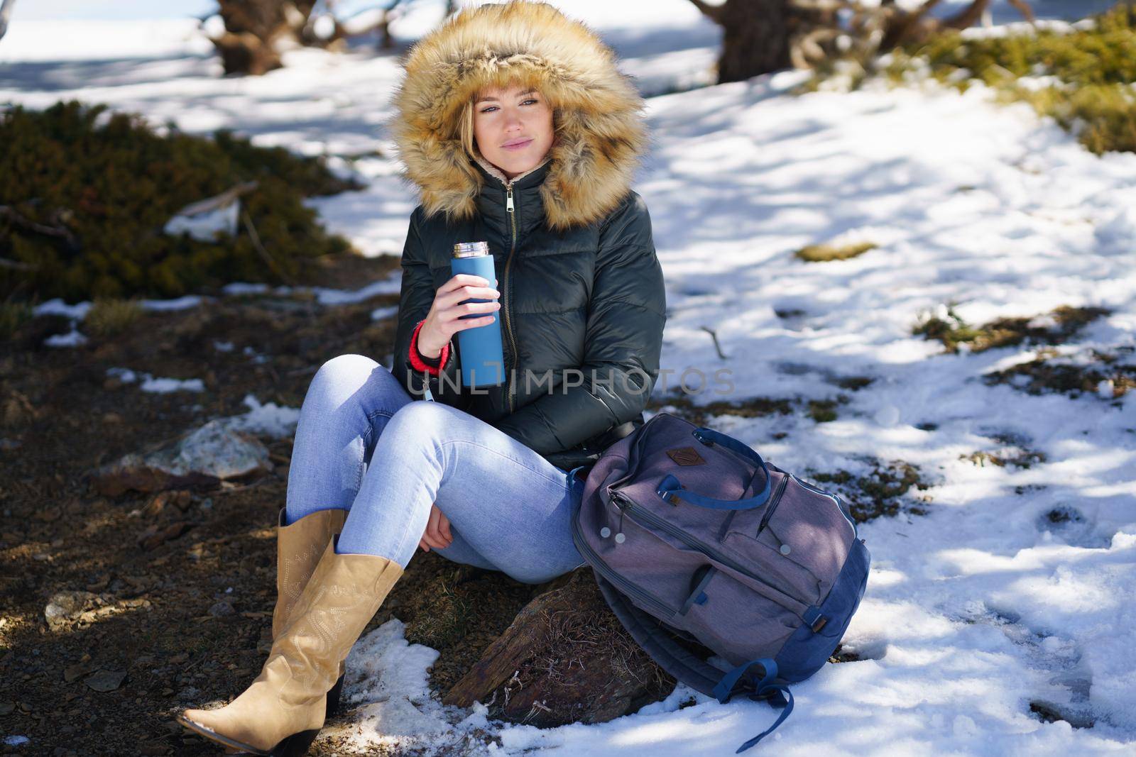 Young woman drinking something hot from a metal thermos bottle sitting on a rock in the snowy mountains., in Sierra Nevada, Granada, Spain. Female wearing winter clothes.