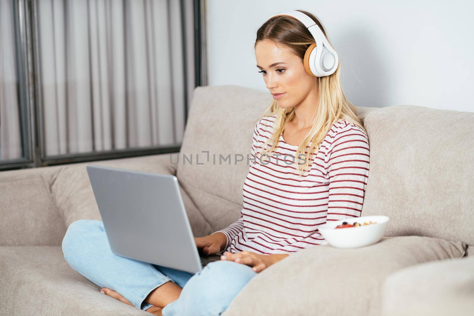 Young blonde woman with headphones and laptop on the sofa at home.