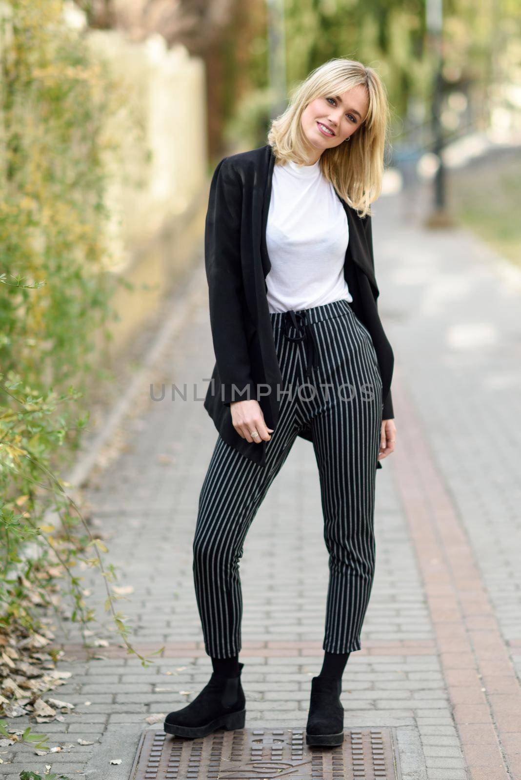 Pretty blonde woman smiling in urban background. Young girl wearing black blazer jacket and striped trousers standing in the street. Pretty female with straight hair hairstyle and blue eyes.
