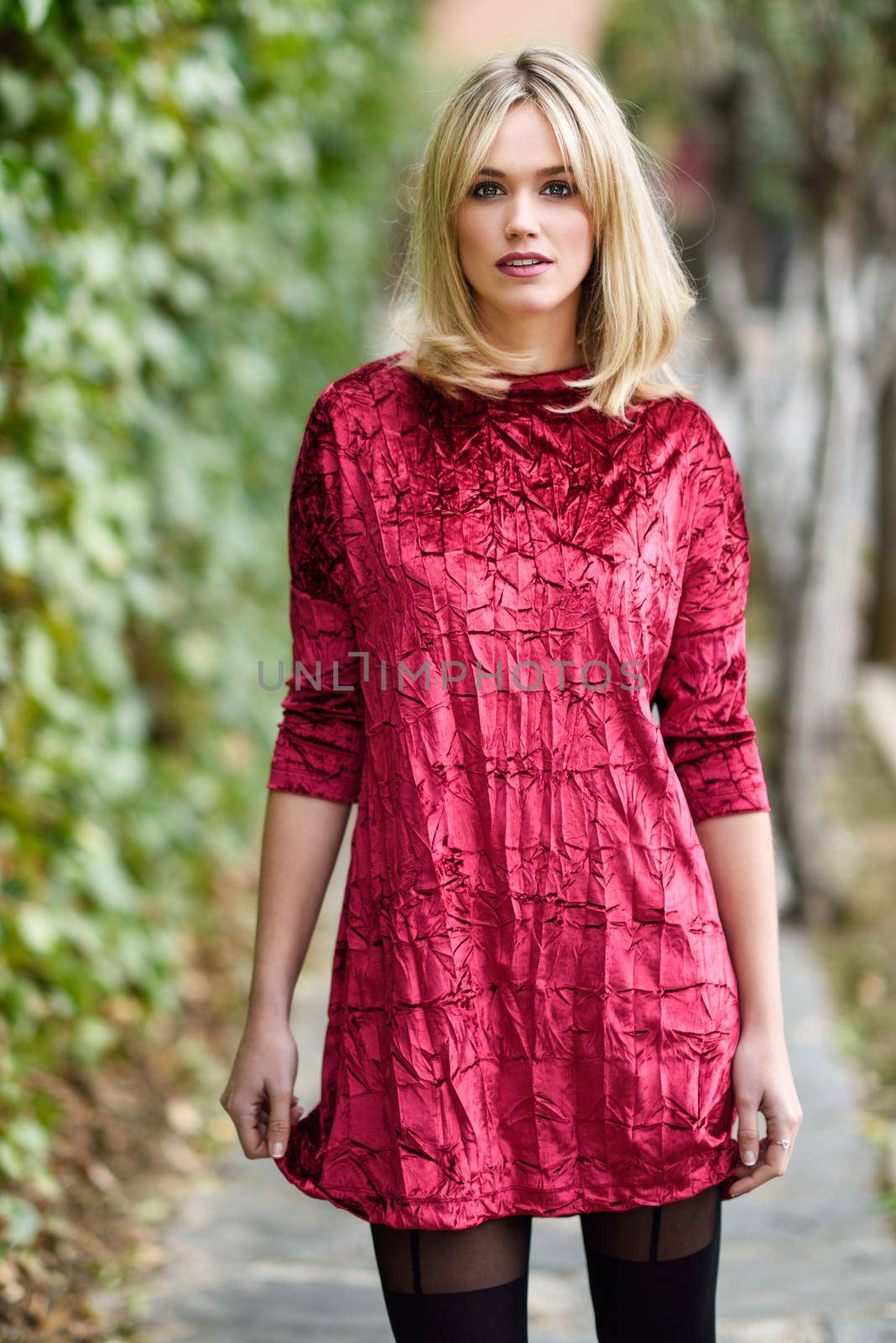 Beautiful blonde woman in green leaves background. Young girl wearing red dress standing in the street. Pretty female with frizzy hairstyle and blue eyes.