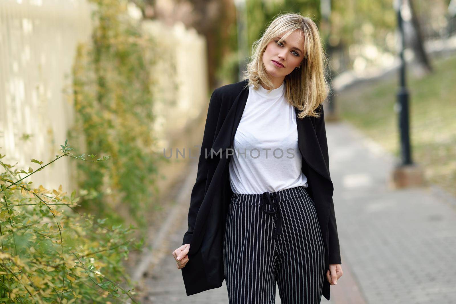 Beautiful blonde woman in urban background. Young girl wearing black blazer jacket and striped trousers standing in the street. Pretty female with straight hair hairstyle and blue eyes.