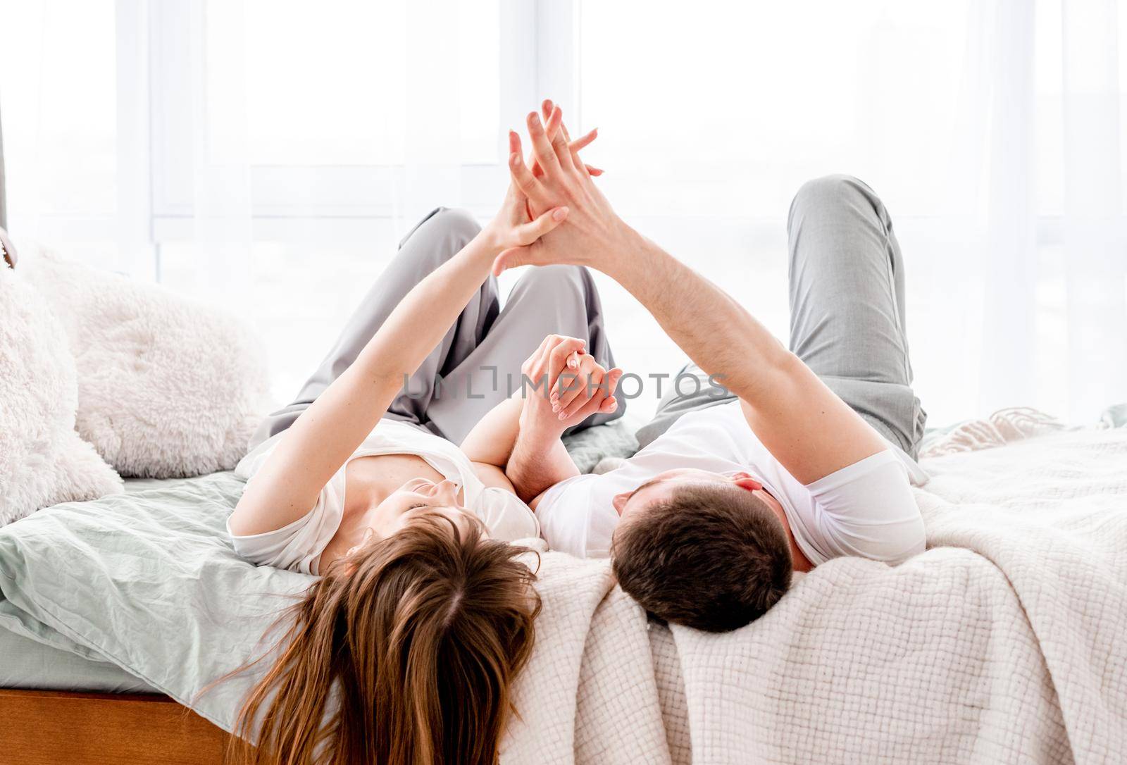 Beautiful young couple lying in the bed and hold hands of echa other in sunny room with panoramic window. Girl and guy wearing pajamas spend time together in the bedroom. Lovely family morning