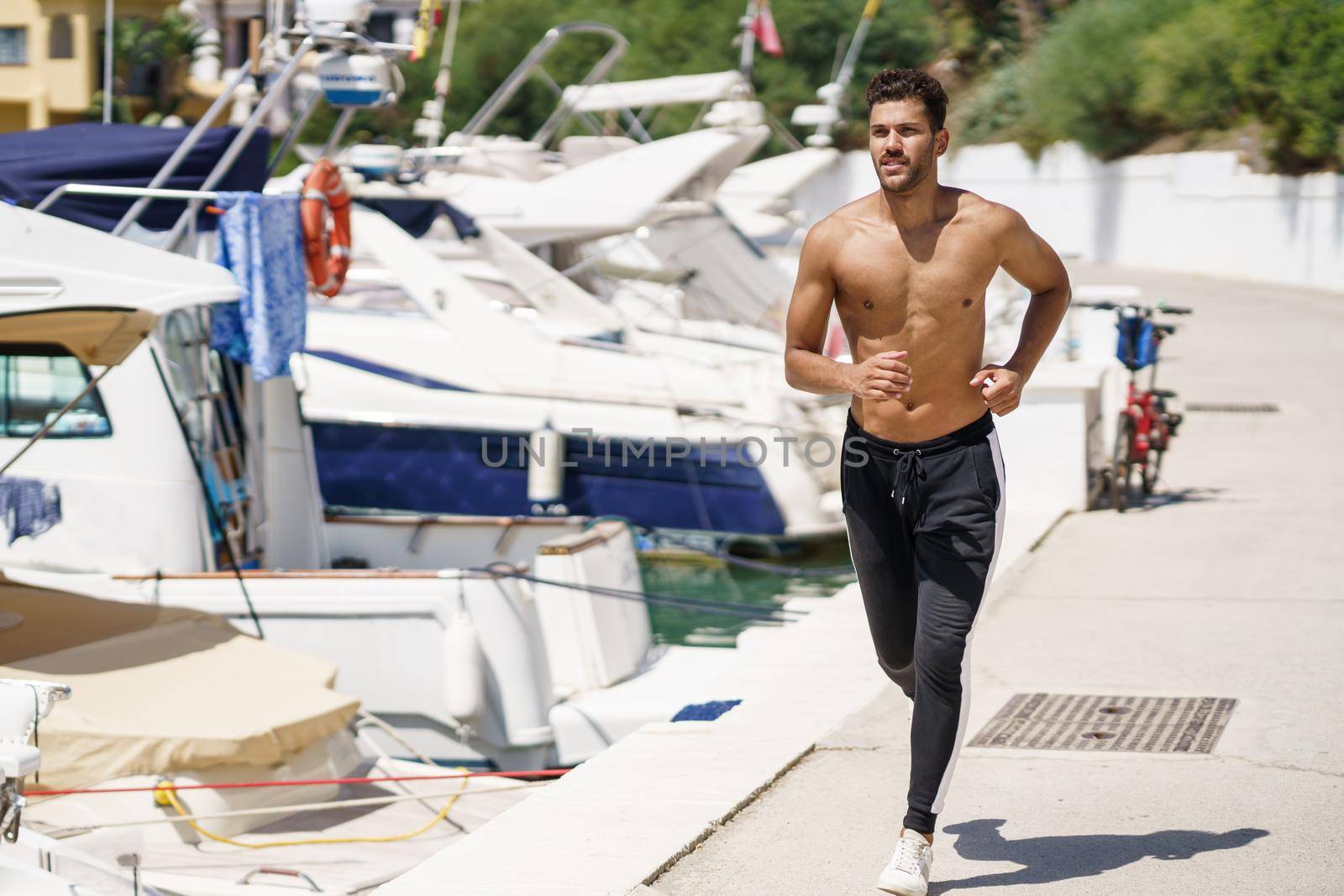 Young man with an athletic body running shirtless through a harbour full of boats.