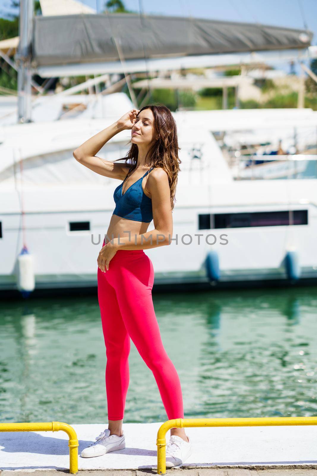 Fitness girl, model of fashion, in red sportswear outfit posing on waterfront harbour.