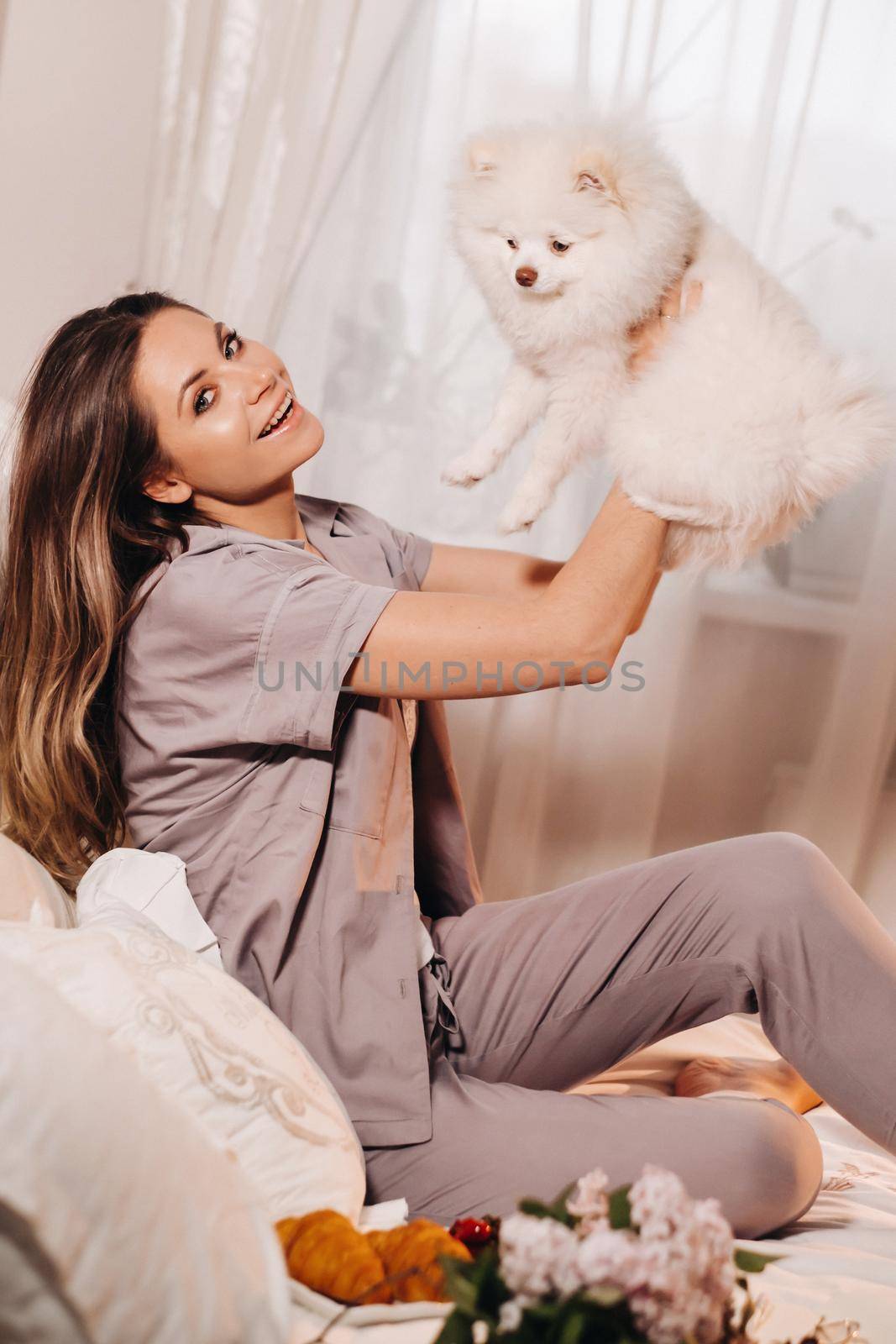 a girl in pajamas sits in bed at night with her white dog watching a laptop and eating sweets.Girl with a dog Spitzer at home in bed by Lobachad