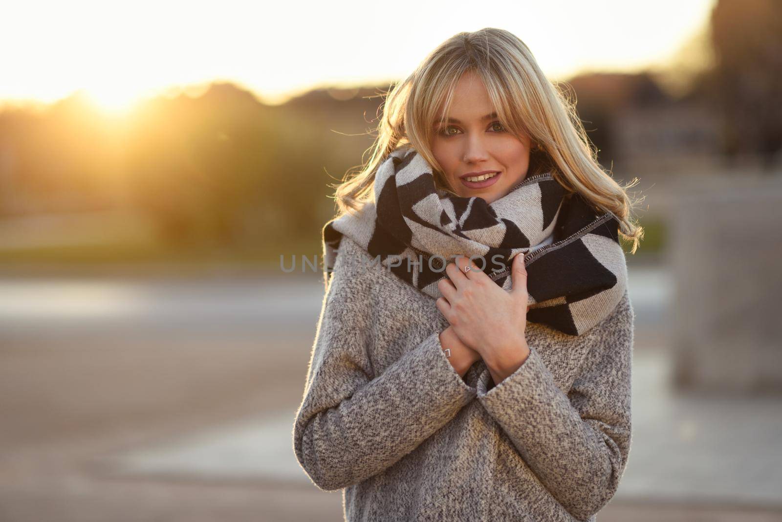 Attractive blonde woman in urban background with sun backlight. Young girl wearing winter coat and scarf standing in the street. Pretty female with straight hair hairstyle and blue eyes.