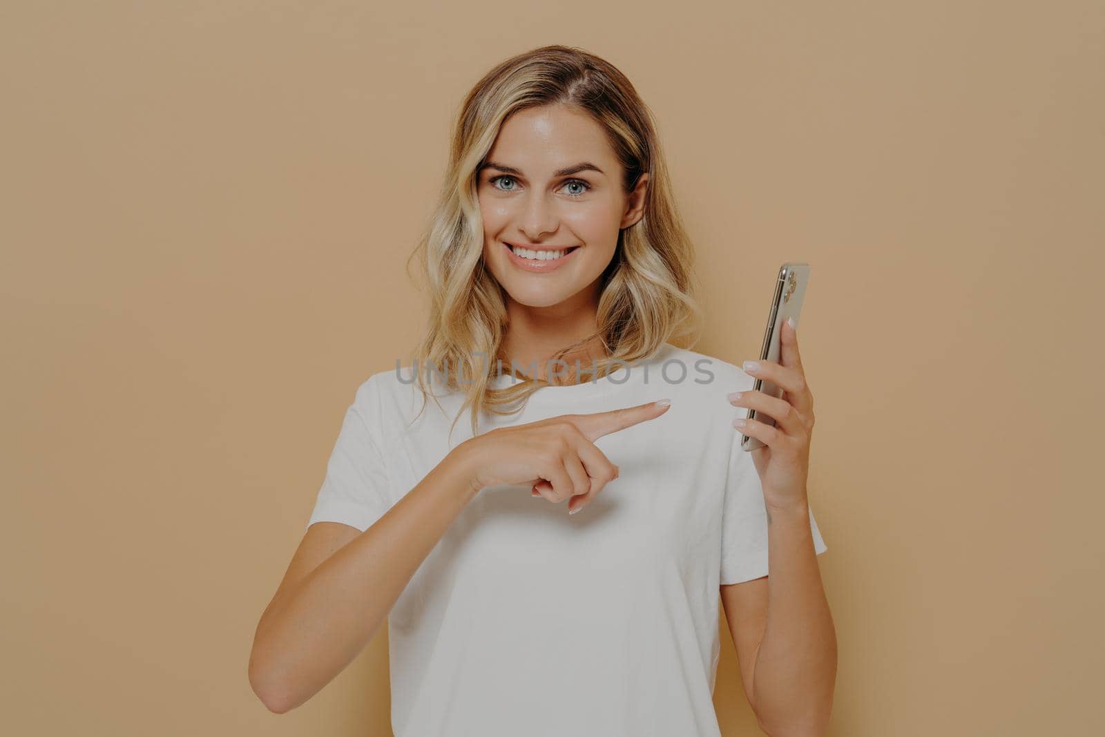 Cute excited female pointing on smartphone with forefinger and looking at camera with broad smile, reading good news in internet and feeling happy while posing isolated over beige background