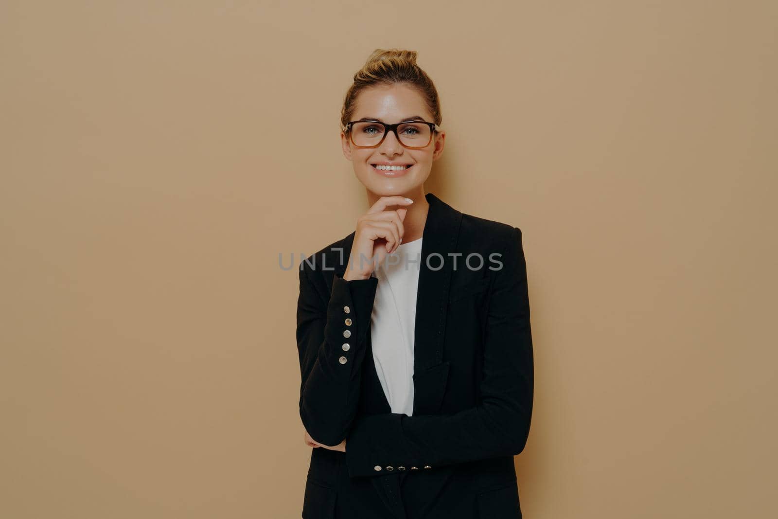 Confident european young female in spectacles and black blazer over white tshirt touching her chin tenderly with hand and smiling widely at camera, expressing positive emotions while posing in studio