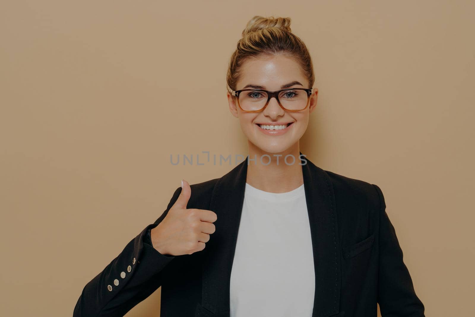 Portrait of positive woman office worker in spectacles and formal outfit showing thumb up and looking at camera with wide smile, being happy with work done, standing isolated over beige background