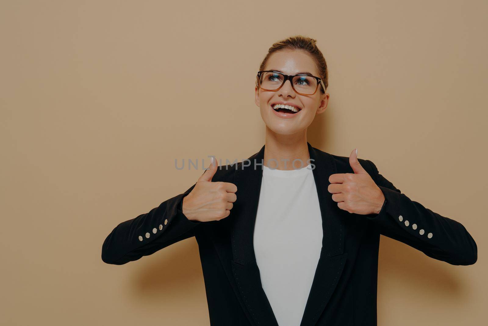 Portrait of positive female customer in eyeglasses with broad smile showing thumbs up with both hands and looking at copy space with happy expression while posing isolated over beige background