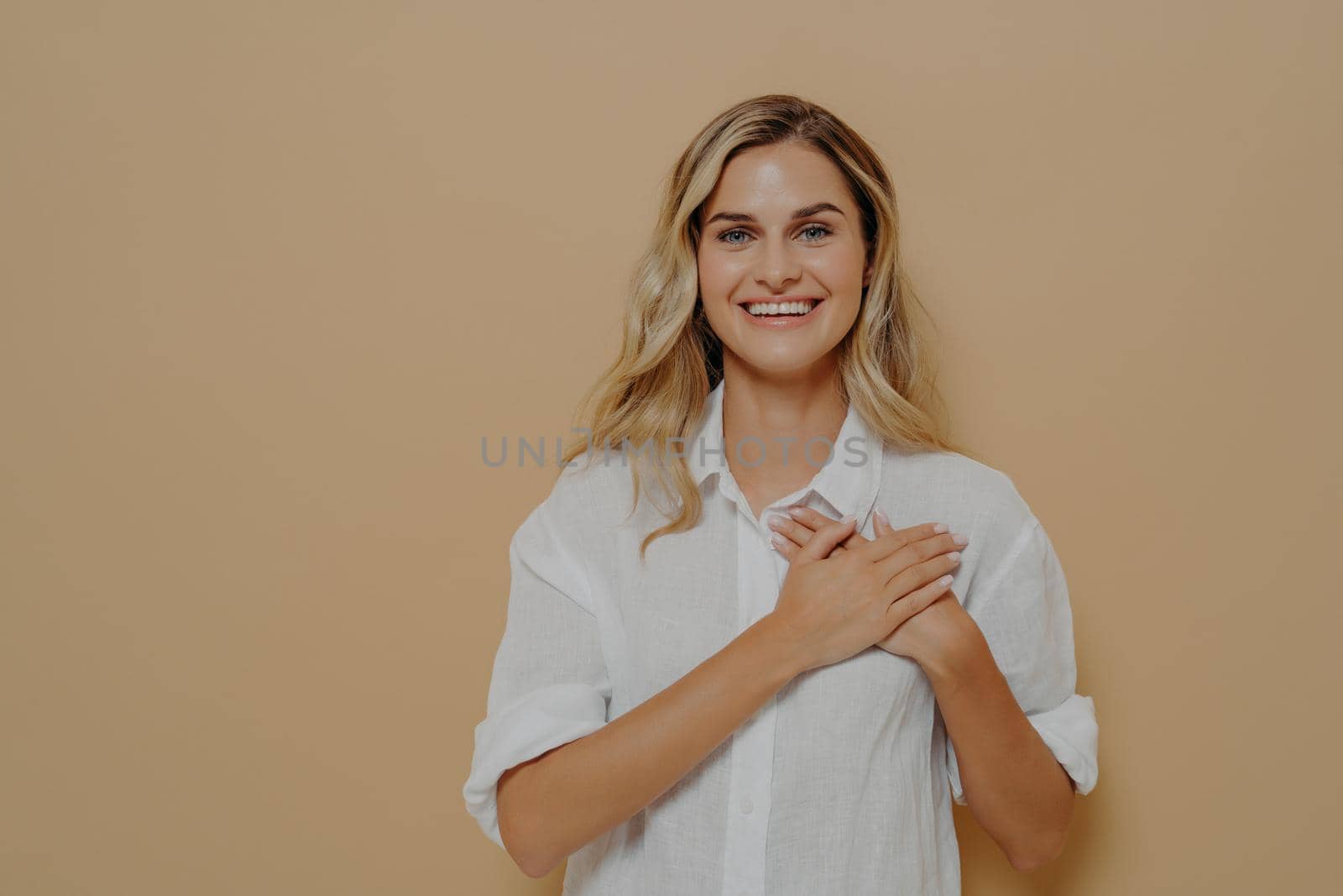 Portrait of admiring cute woman in love with blond hair holding hands on chest sighing with romantic smile, gazing at camera as feeling grateful and appreciative isolated on beige background