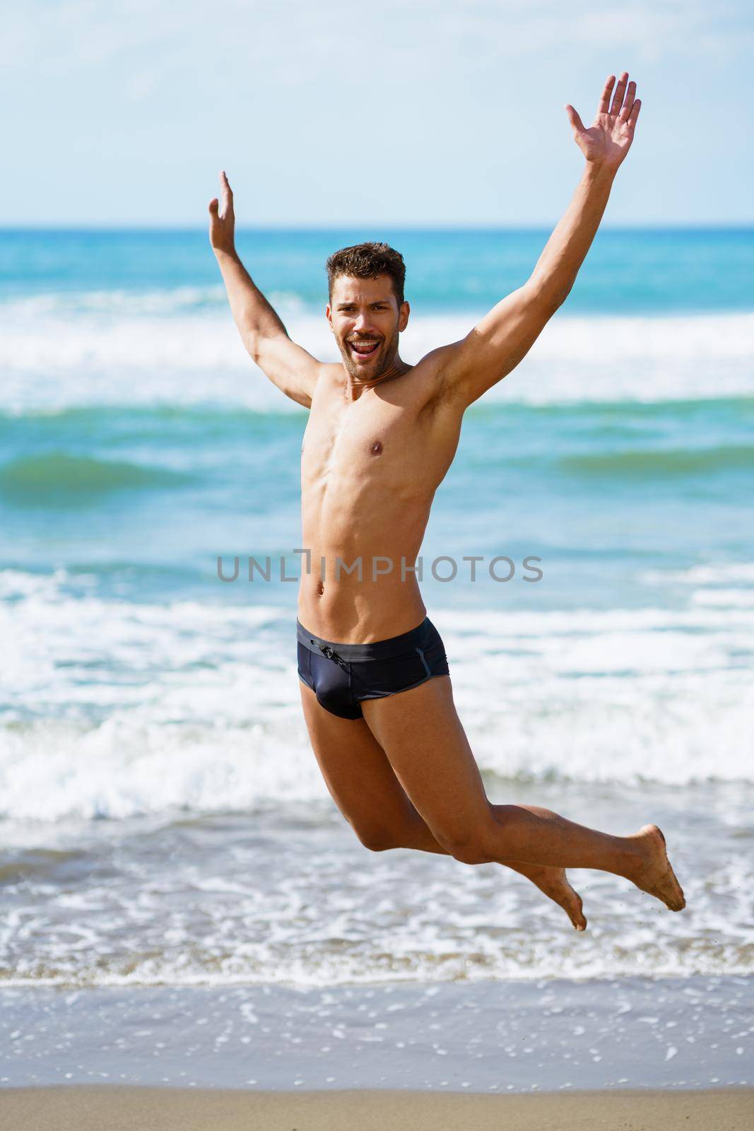 Funny young man with beautiful body in swimwear jumping on a tropical beach.
