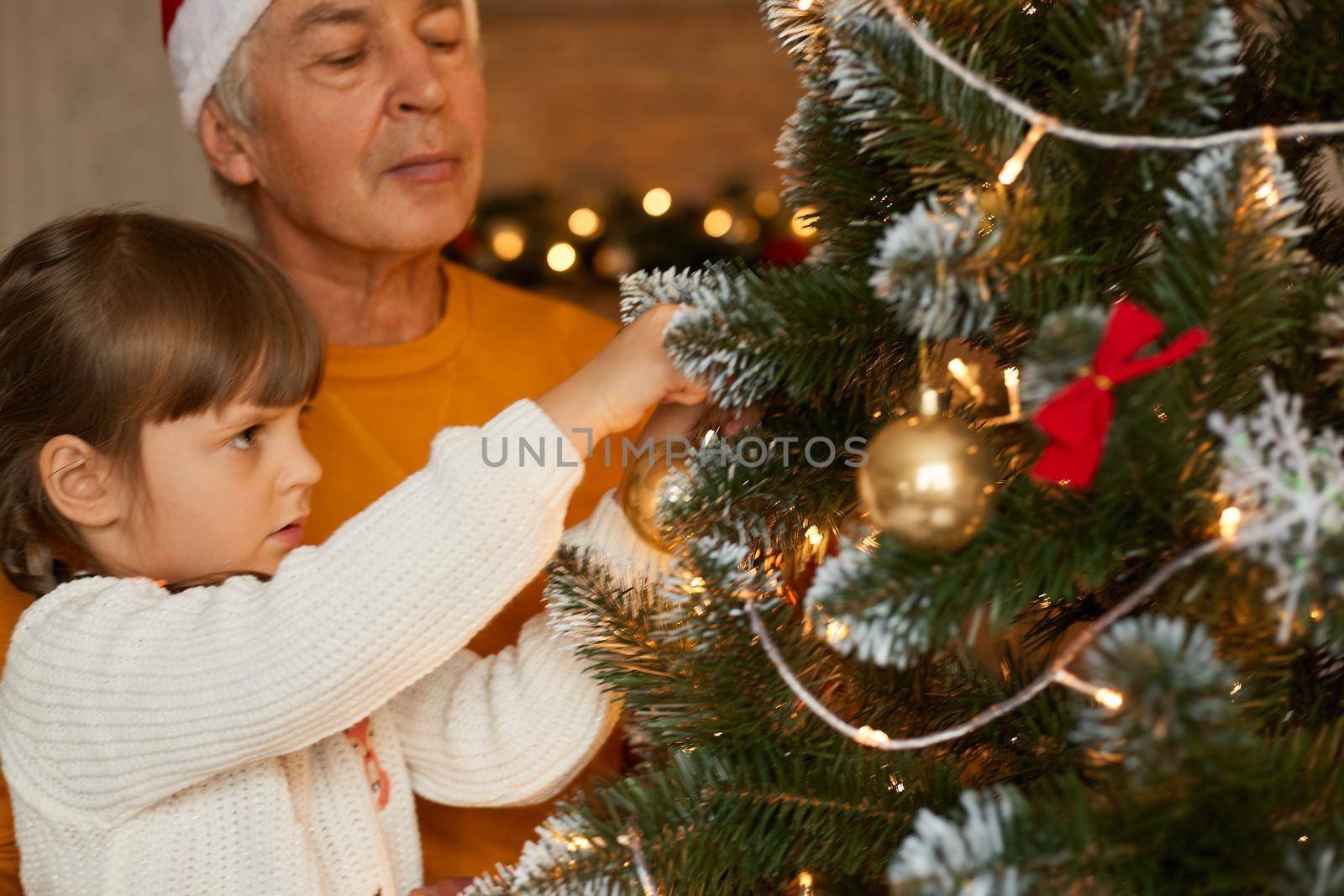 Happy family decorating xmas tree, grandfather wearing orange sweater and little girl in white jumper posing in living room, beautiful xmas tree with bubbles and bows. by sementsovalesia