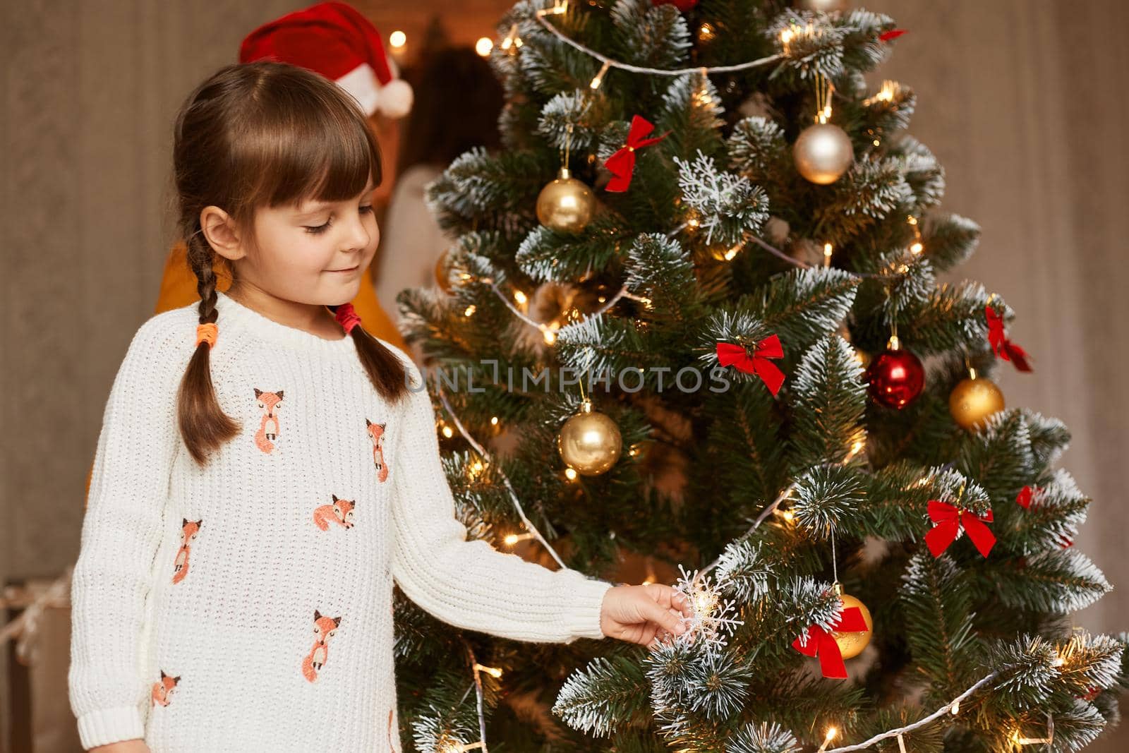 Adorable little girl decorating a Christmas tree with colorful glass baubles at home, female kid with pigtails wearing white sweater posing at home during new year eve.