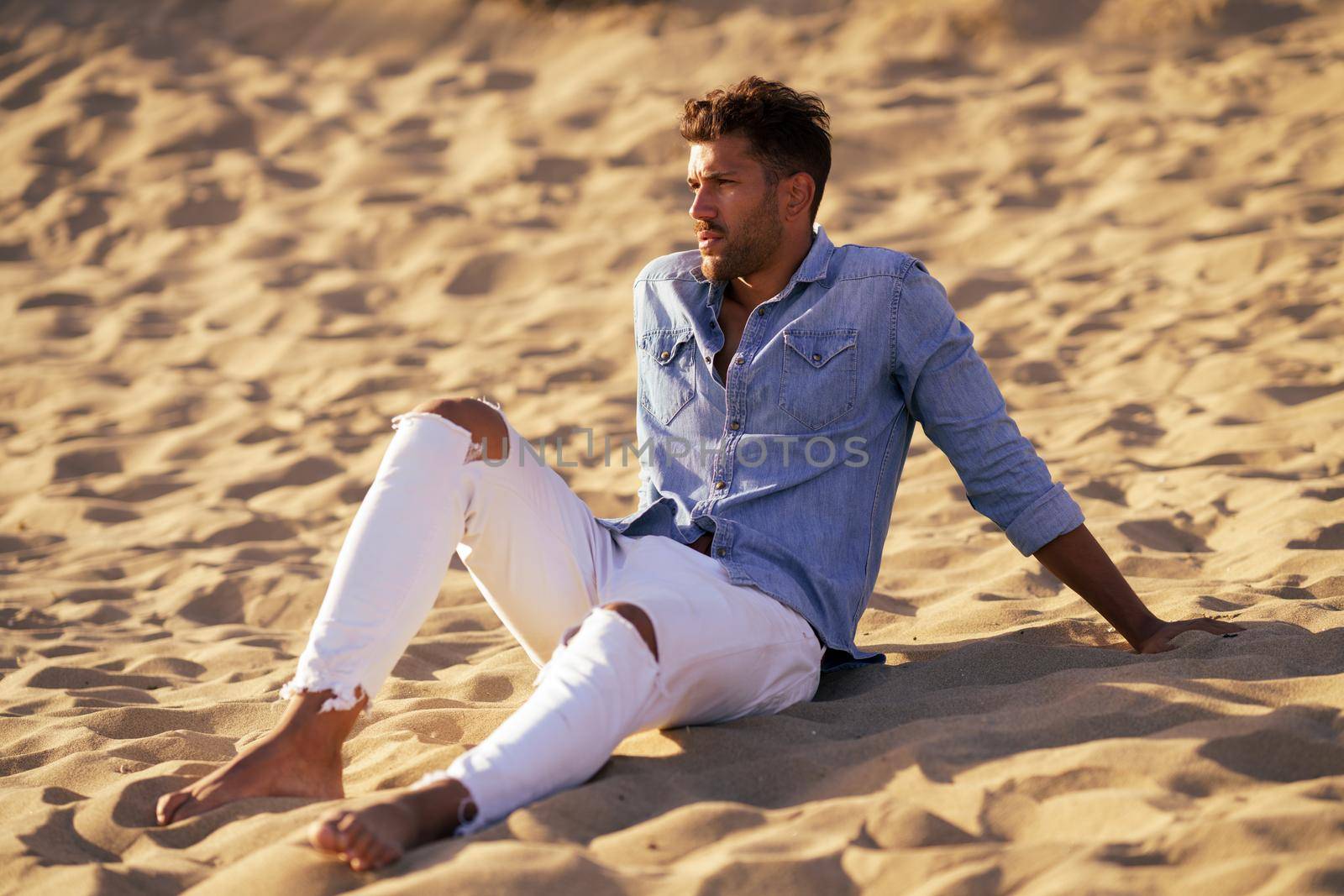 Attractive man sitting on the sand of the beach looking at the horizon.