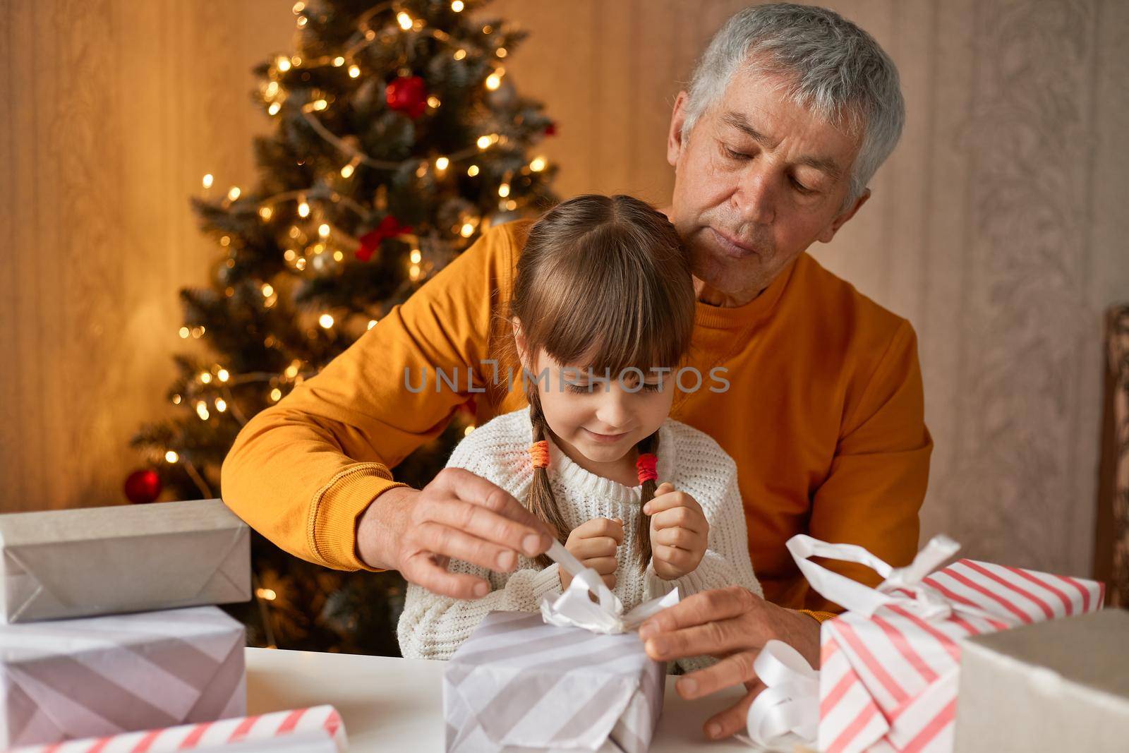 Mature man posing in festive living room with kid, packing Christmas gifts near fir tree, looking concentrated at boxes, child in white casual shirt, mature male in orange jumper.