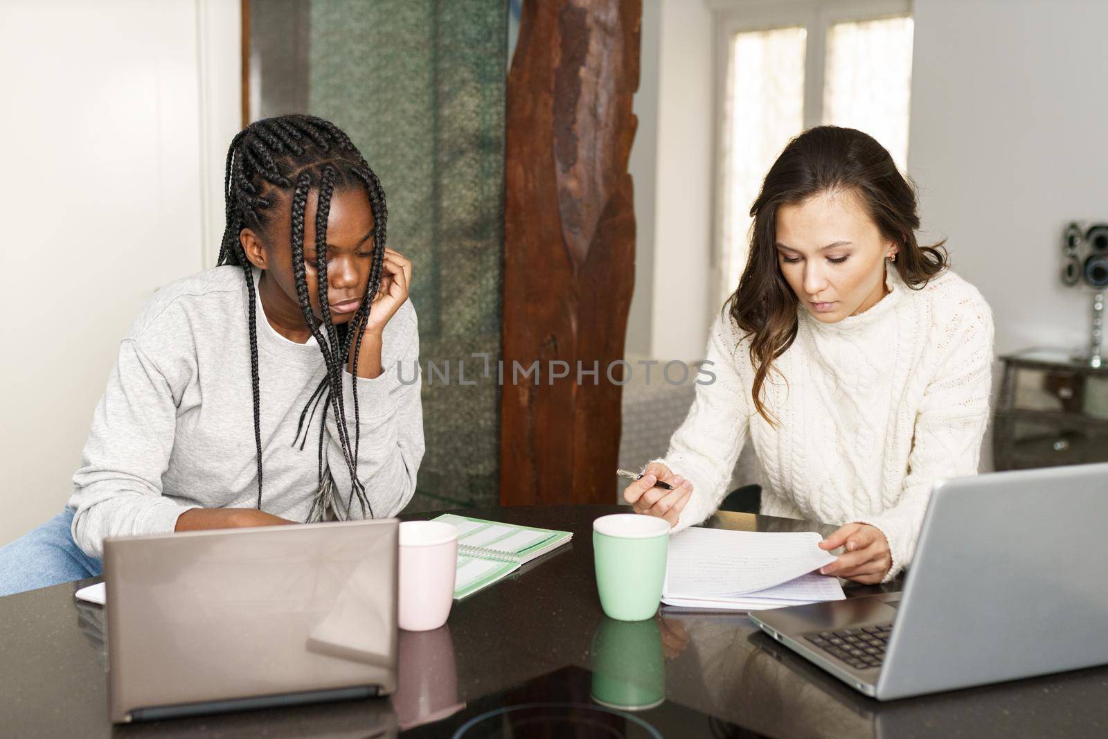 Two college girls studying together at home with laptops while drinking coffee. Multiethnic women.