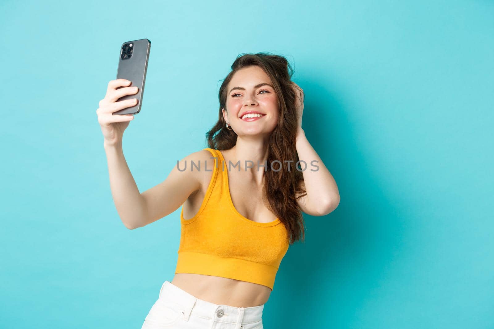 Beautiful modern girl taking selfies on vacation, posing in summer clothes and looking at smartphone camera, standing against blue background by Benzoix