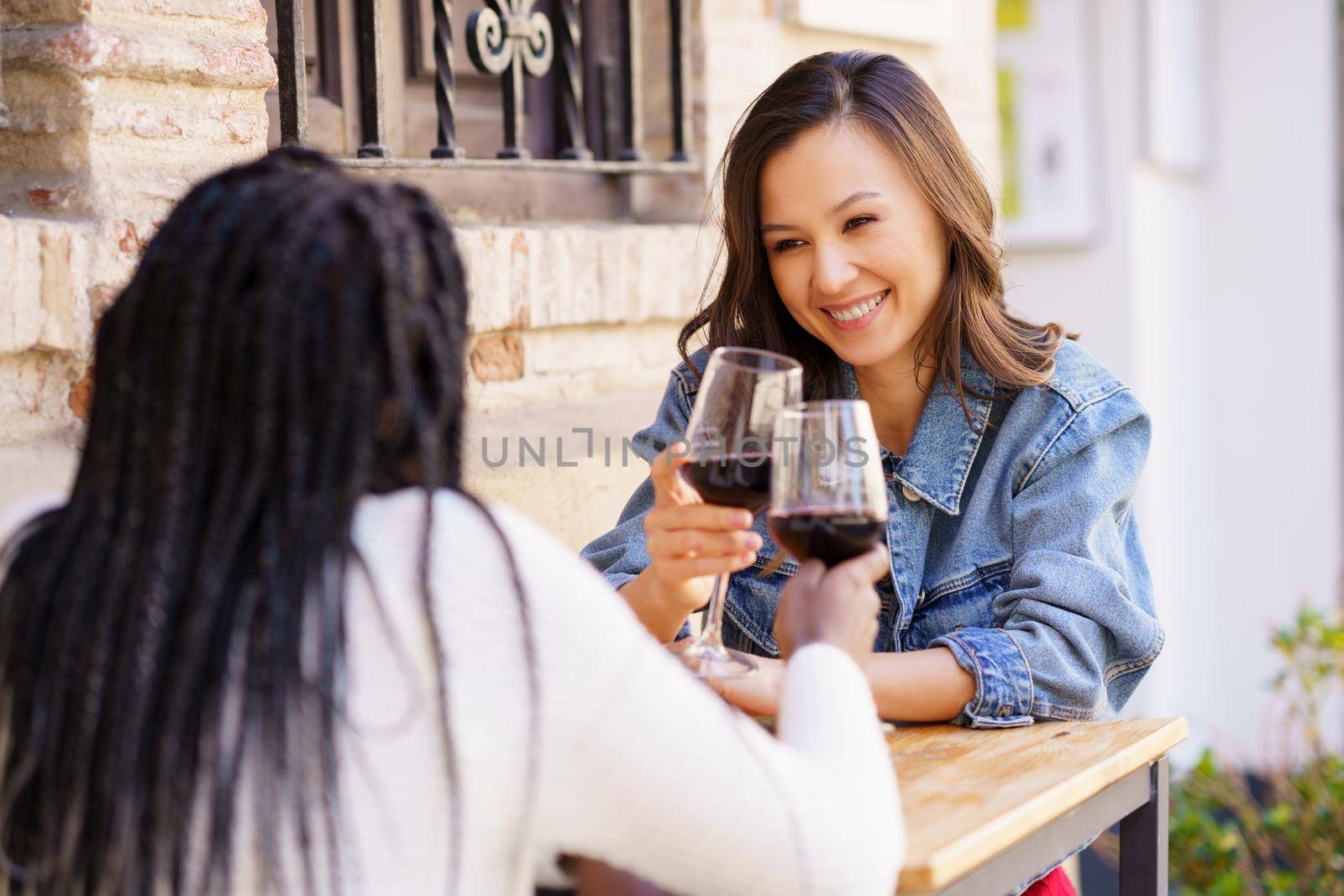 Two women making a toast with red wine sitting at a table outside a bar. by javiindy