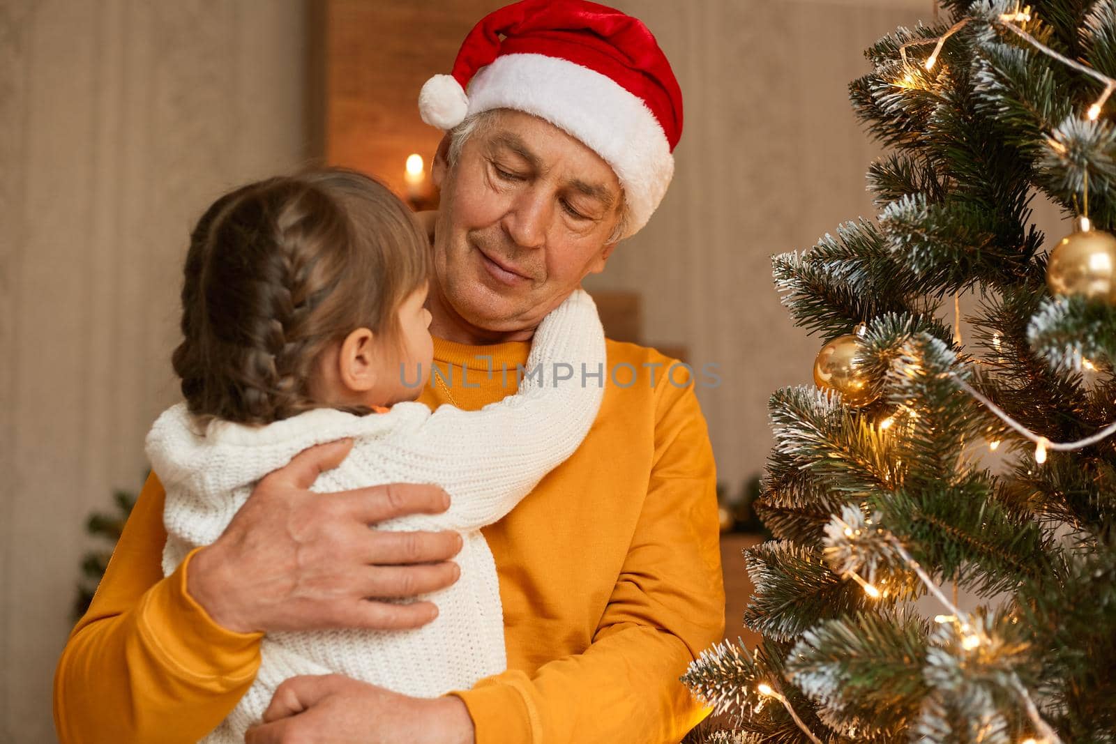 Happy family on Christmas eve posing in festive room, mature man hugging granddaughter in white sweater and with pigtails, family expressing love, celebrating winter holiday together.
