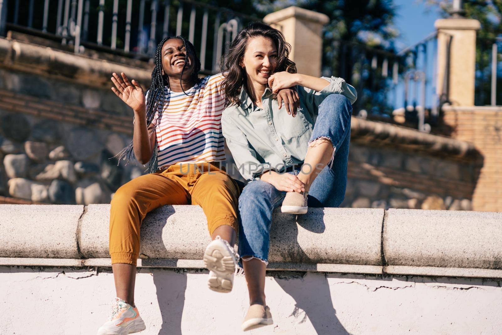 Two friends having fun together on the street sitting on a urban wall. Multiethnic women.