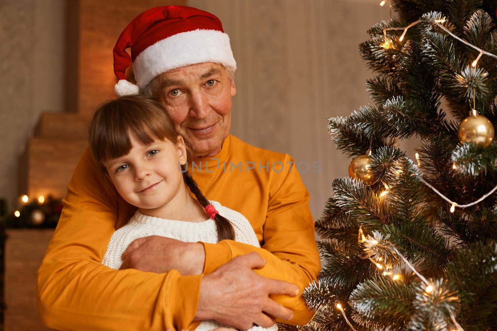 Cute child sitting with grandpa looking at camera with happy expression, grandfather hugging little girl, posing in room with festive new year decoration.