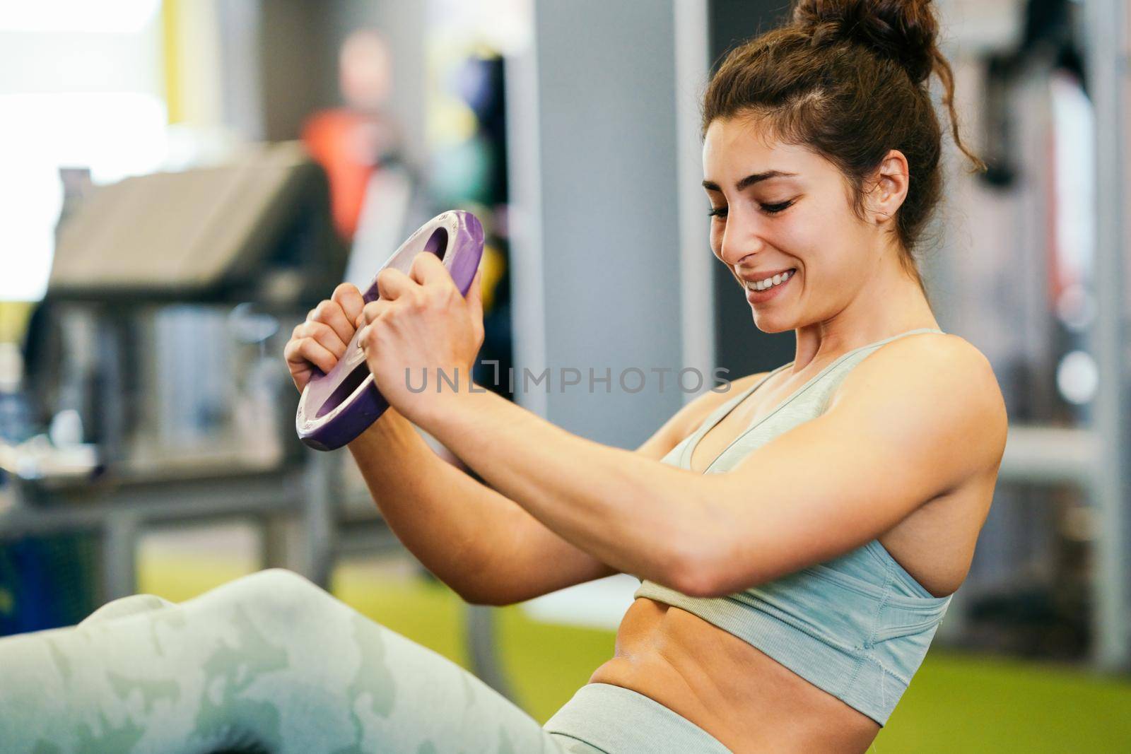 Sporty young female, wearing sportswear, on yoga mat doing situps with plate weights in gym. Fitness woman doing abs crunches.