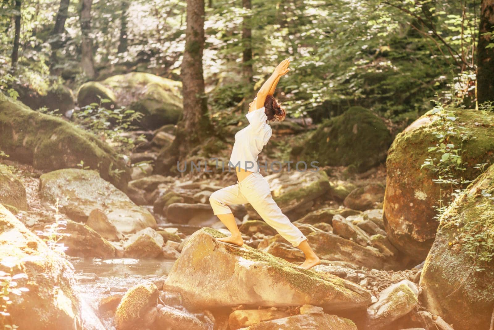 Young woman practicing yoga in pose of warrior on stone in summer forest outdoor. Image with sunlight effect.