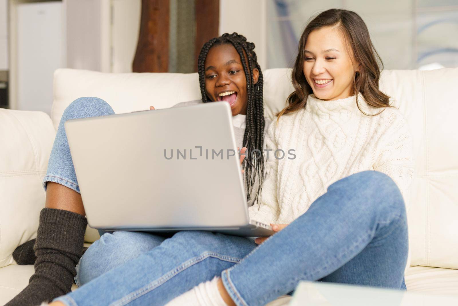 Two female student friends sitting on the couch at home using a laptop. Multiethnic women.