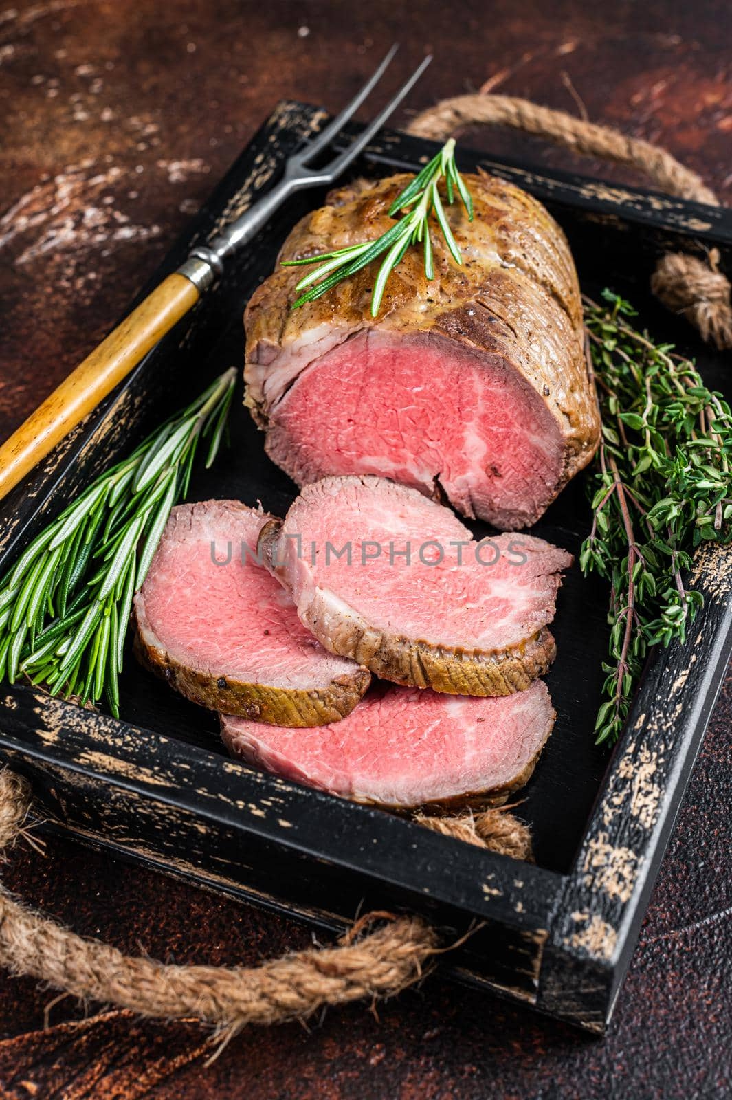 Roast beef round fillet meat in a wooden tray with herbs. Dark background. Top view.