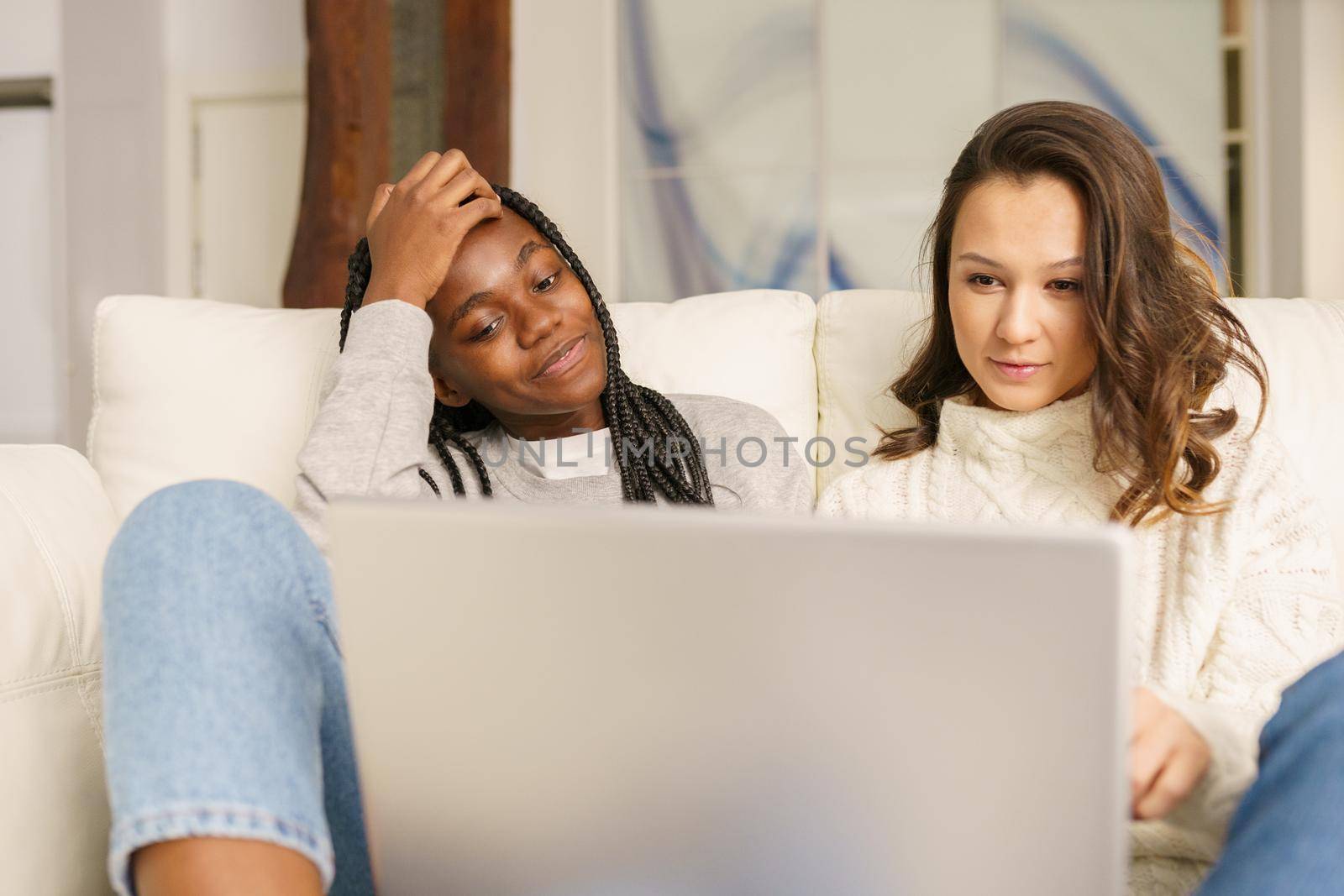 Two female student friends sitting on the couch at home using a laptop. Multiethnic women.
