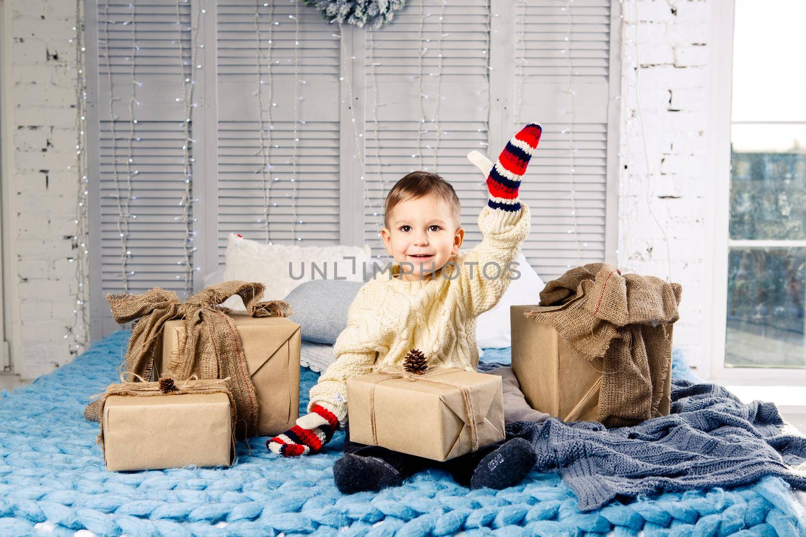 little funny playful boy a child sits on a bed on Christmas day with gift boxes in white wool knitted sweater and big bright mittens on it and laughs out loud. In interior there is a festive decor by Tomashevska