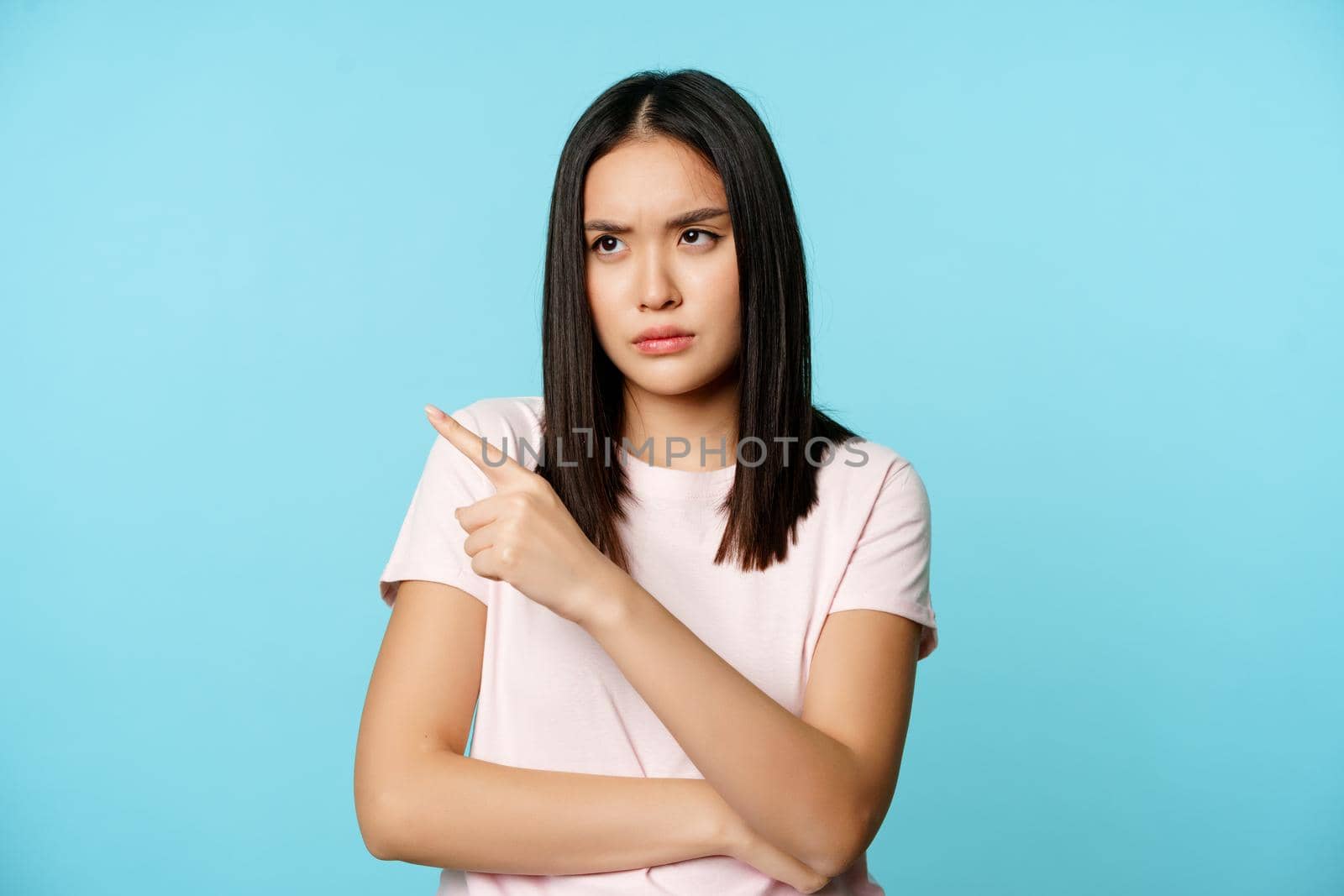 Angry asian woman sulking, frowning upset, pointing fingers at upper left corner, standing in t-shirt over blue background.