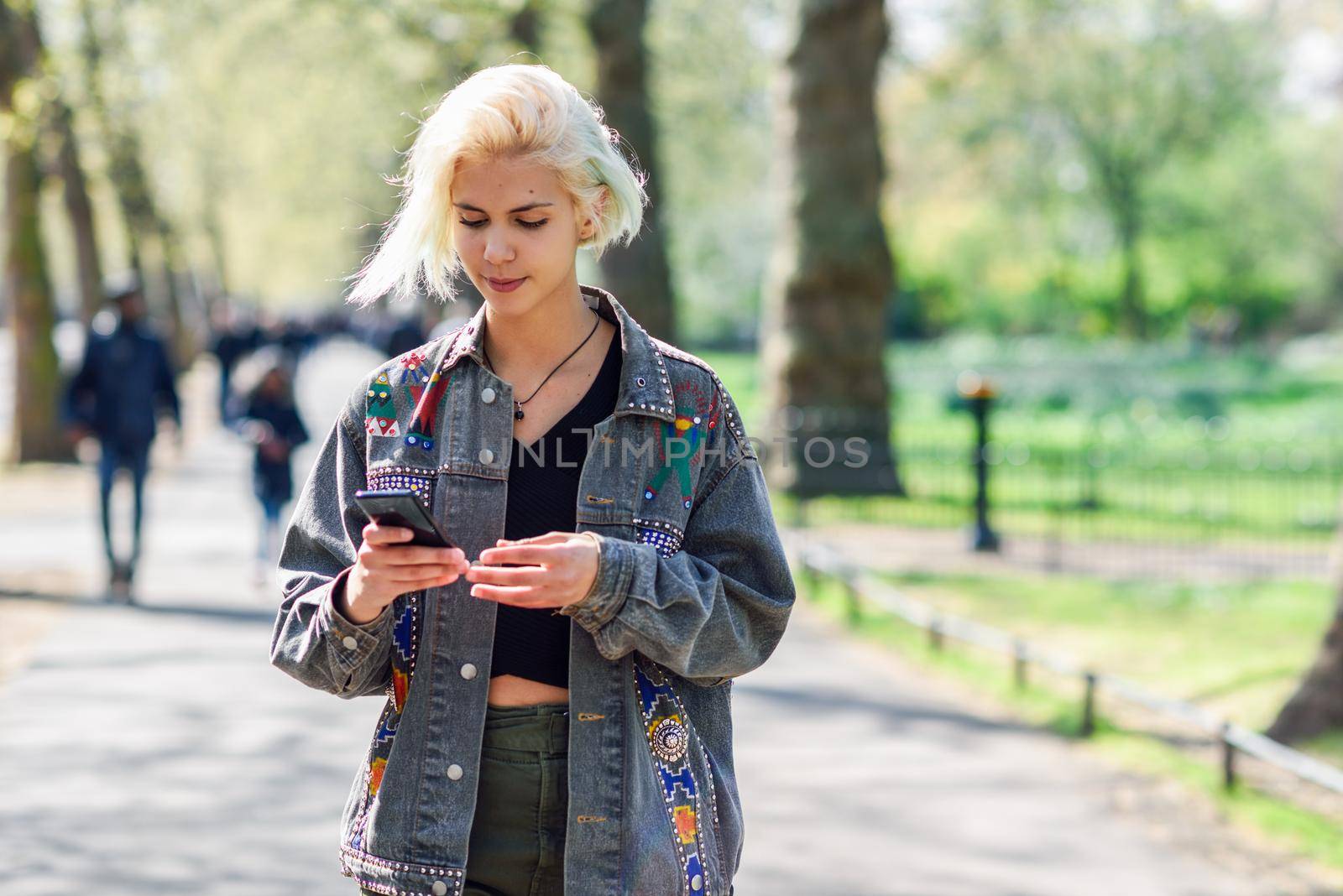 Young urban woman with modern hairstyle using smartphone walking in street in an urban park. by javiindy