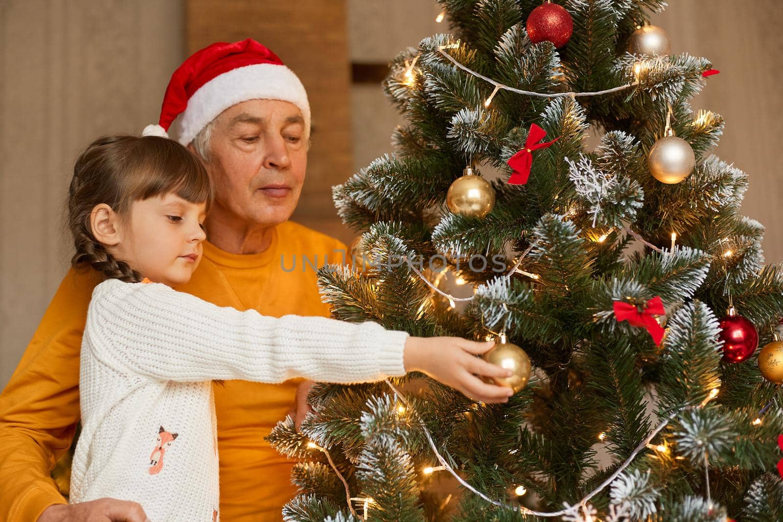 Senior man with his little granddaughter decorating Christmas tree at home, old male wearing santa claus hat and orange shirt, little child in white sweater.