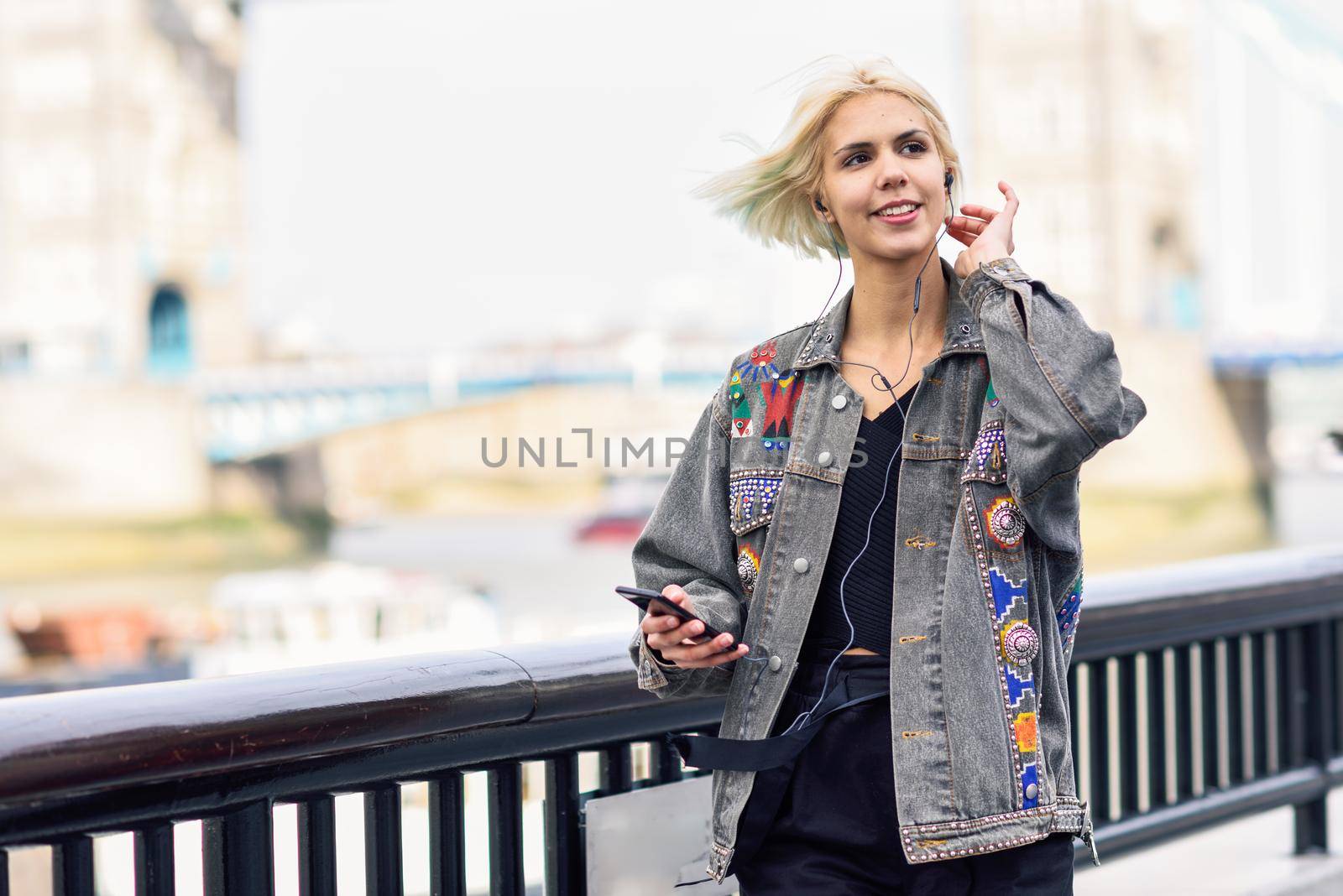 Young woman listening to the music with earphones and smart phon in urban background. Thames River in London.