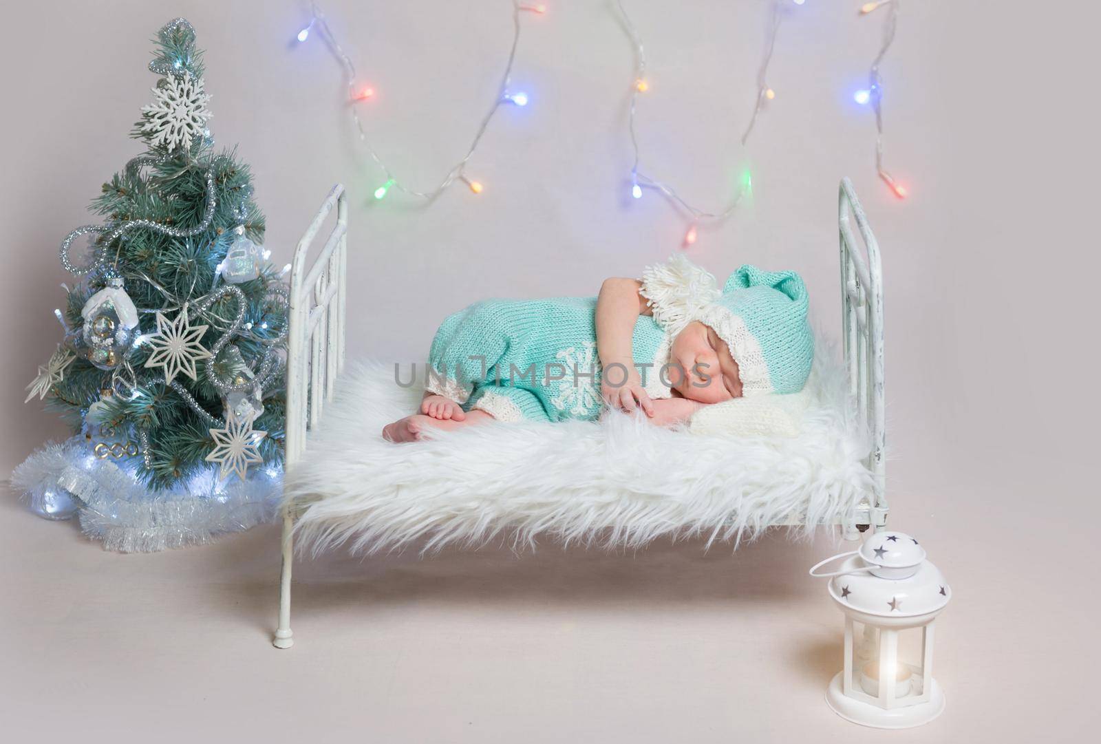 Newborn boy in blue costume sleeping on small cot with Christmas decoration around it. Colourful girlands on the background.