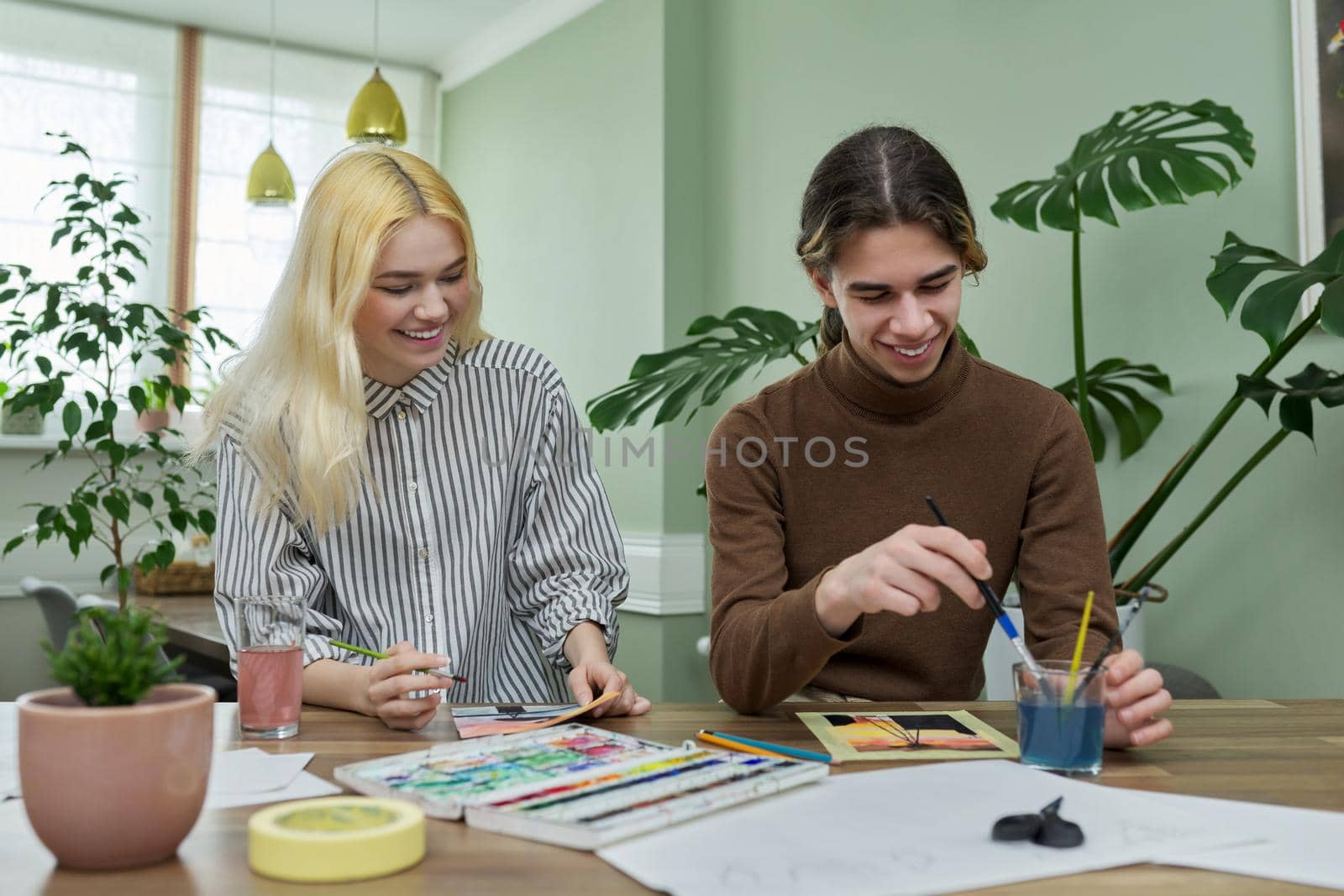 Teenagers painting with watercolors. Couple of guy and girl sitting at table with paints, brushes, drawings. Happy teens talking laughing creative. Creativity, hobby, leisure concept