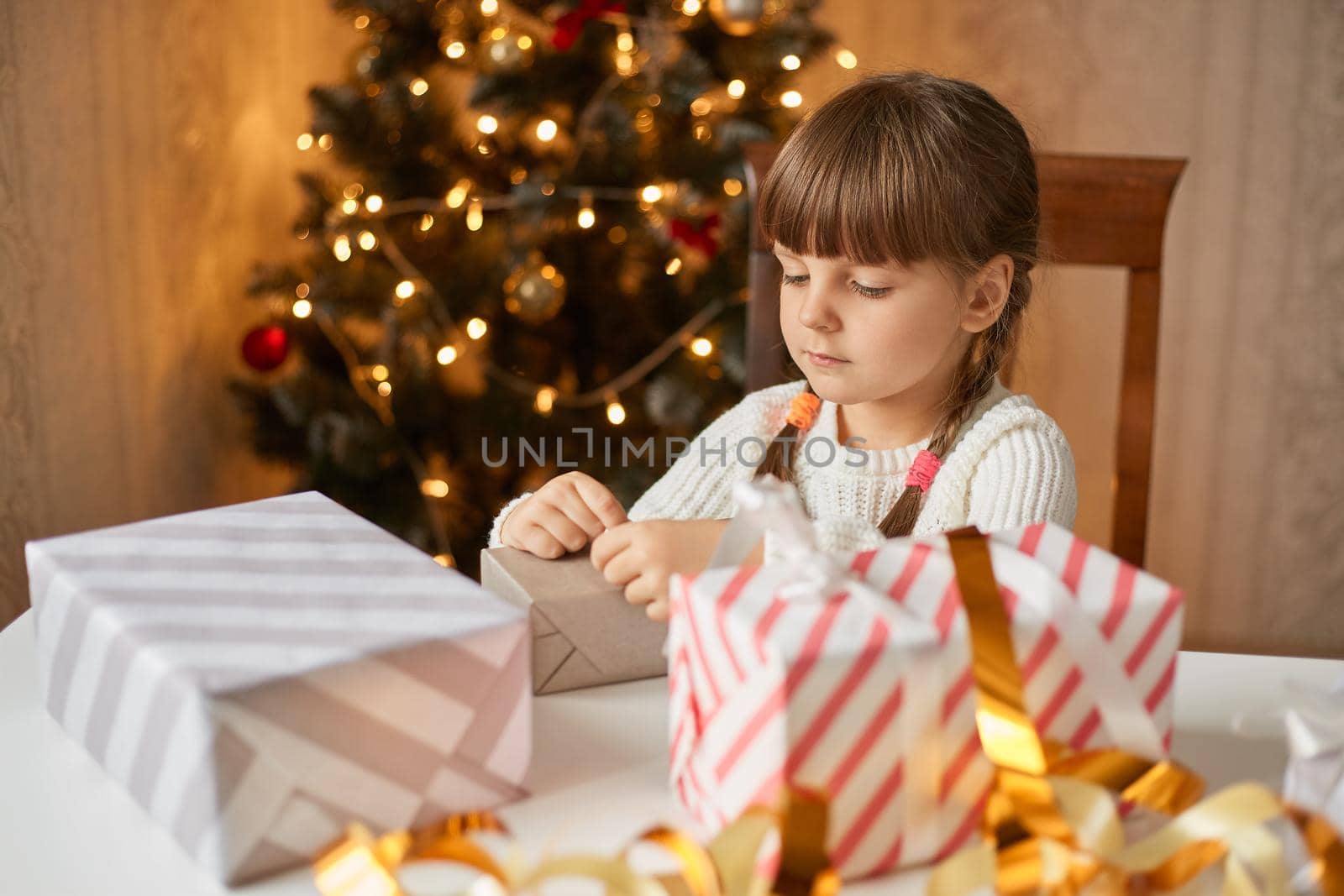 Little concentrated female kid with two pigtails sitting at table and packing christmas box, posing with xmas tree on background, child with concentrated look, making surprise for family.
