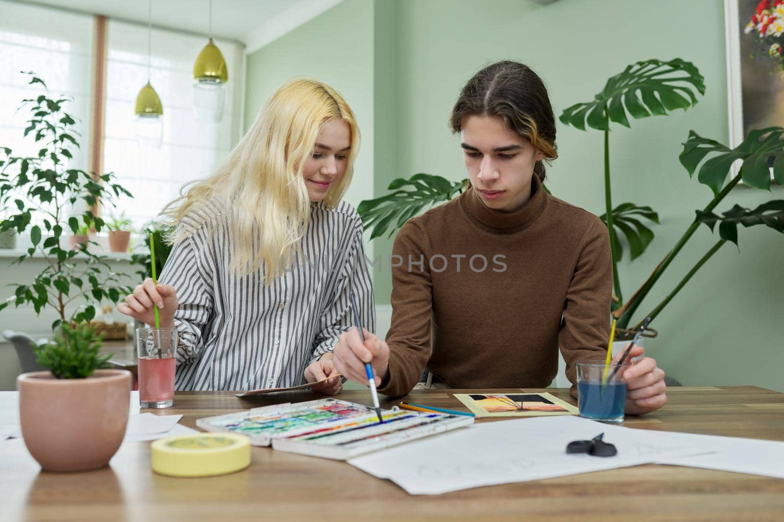 Teenagers painting with watercolors. Couple of guy and girl sitting at table with paints, brushes, drawings. Happy teens talking laughing creative. Creativity, hobby, leisure concept