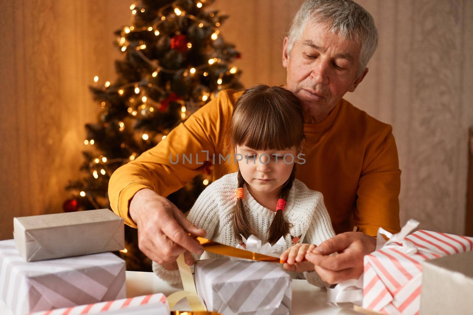 Indoor shot of mature man with his granddaughter sitting at table and preparing for new year eve, packing gift boxes, posing at home near Christmas tree.