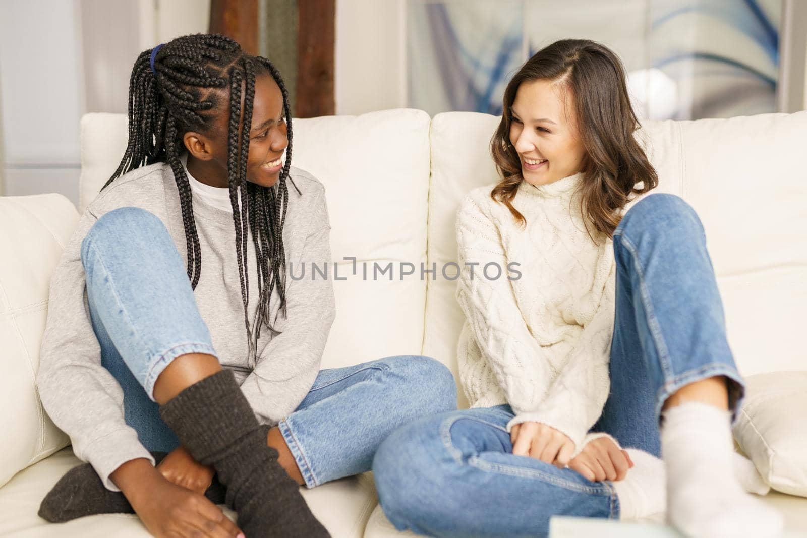 Two female student friends smiling together on the couch at home. Multiethnic women.