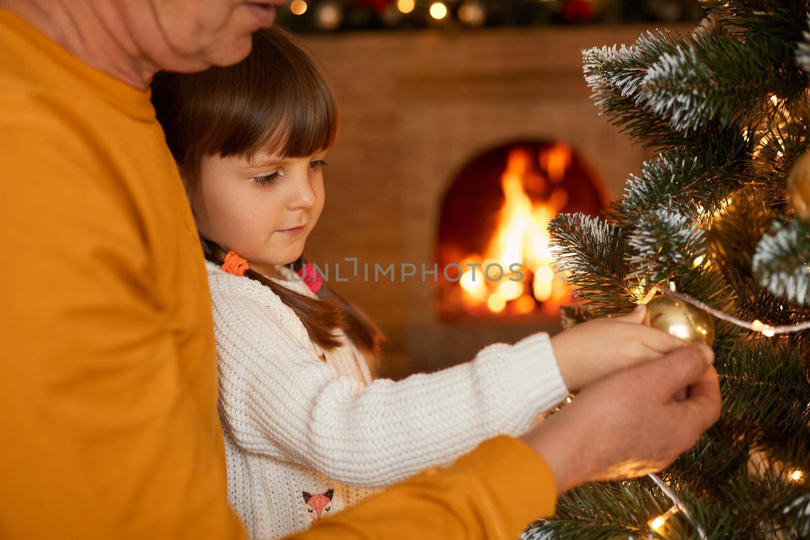 Unknown man with little daughter standing by xmas tree, decorating its with xmas ball, looking at garland's lights, posing with blurred fireplace on background.