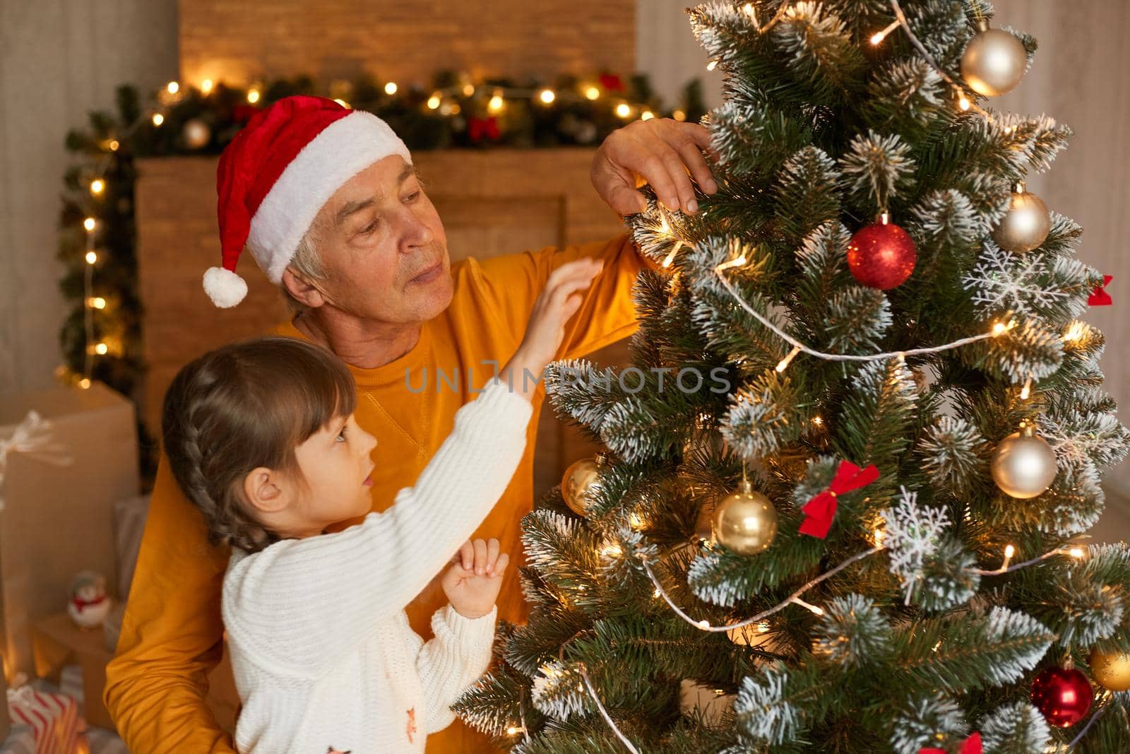 Family decorating christmas tree at home, grandfather spending time with his daughter on new year eve, senior man wearing santa claus hat and yellow shirt, child with pigtails in white jumper. by sementsovalesia