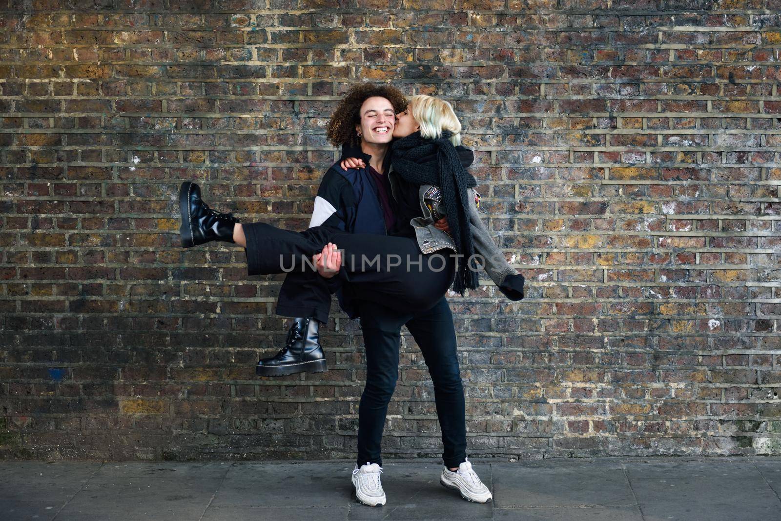 Man holding his girlfriend in his arms in front of a brick wall typical of London by javiindy